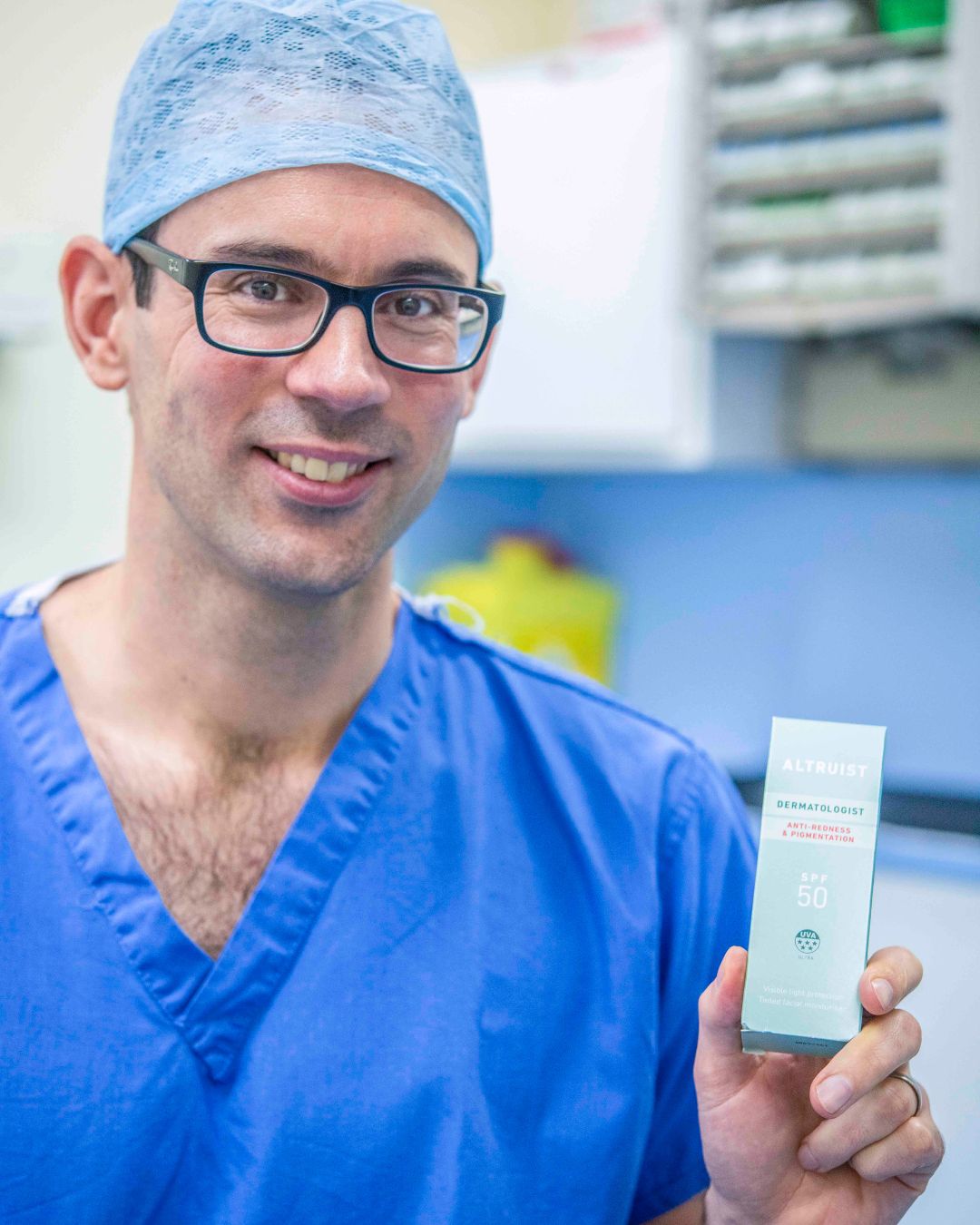 Doctor Andrew Birnie in blue medical scrubs holding an Altruist skincare product in a clinical setting